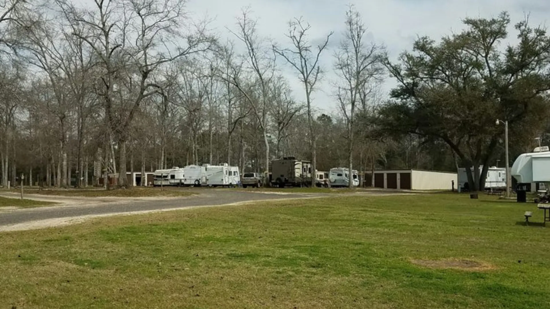 A view of several RVs and travel trailers parked in a grassy, wooded campground setting with storage units visible in the background.