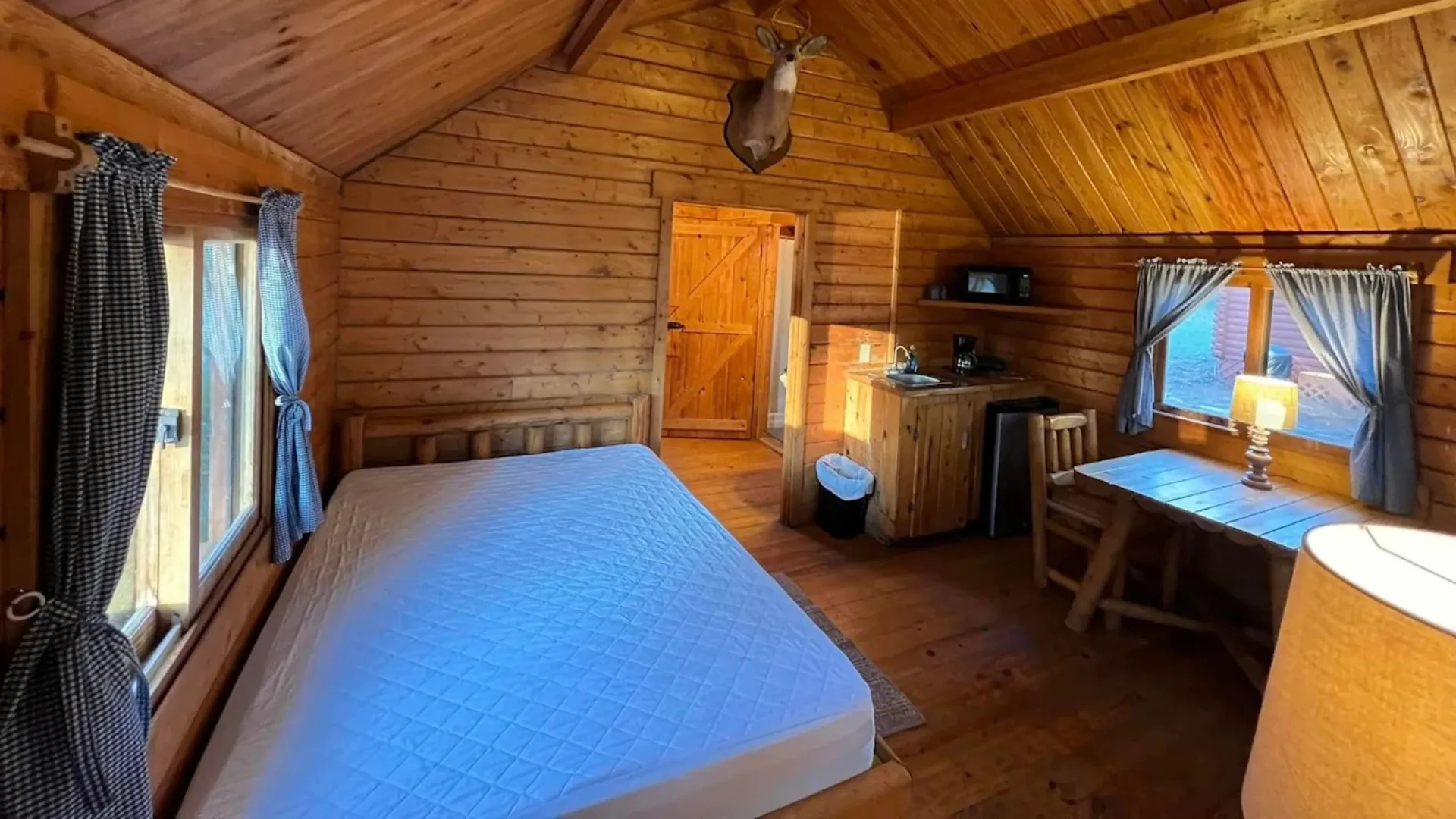 Rustic bedroom inside a Tallahassee RV park cabin rental featuring a wooden bed frame, log-style furniture, a kitchenette, and a mounted deer head on the wall.