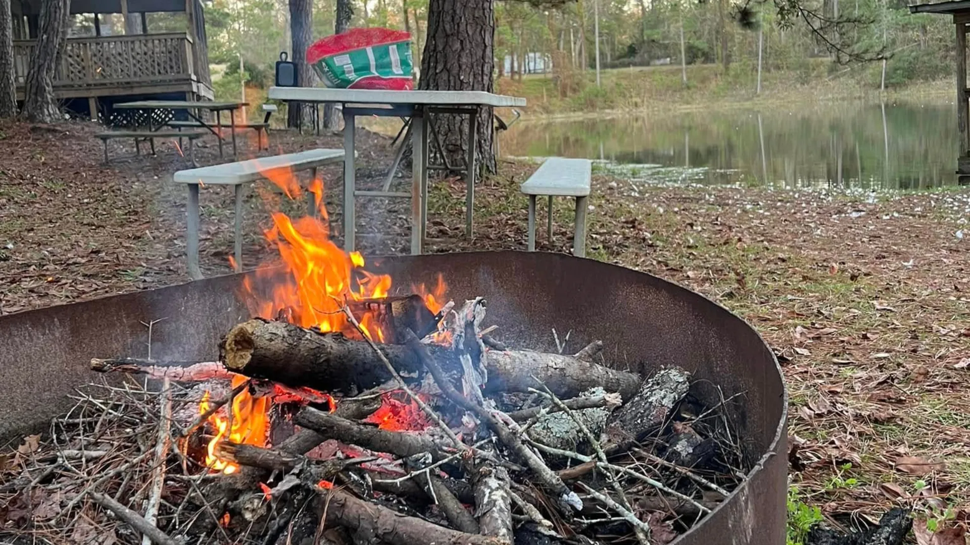 A crackling campfire in a metal fire ring at a lakeside campsite in a Tallahassee RV resort, featuring picnic tables and a scenic water view.