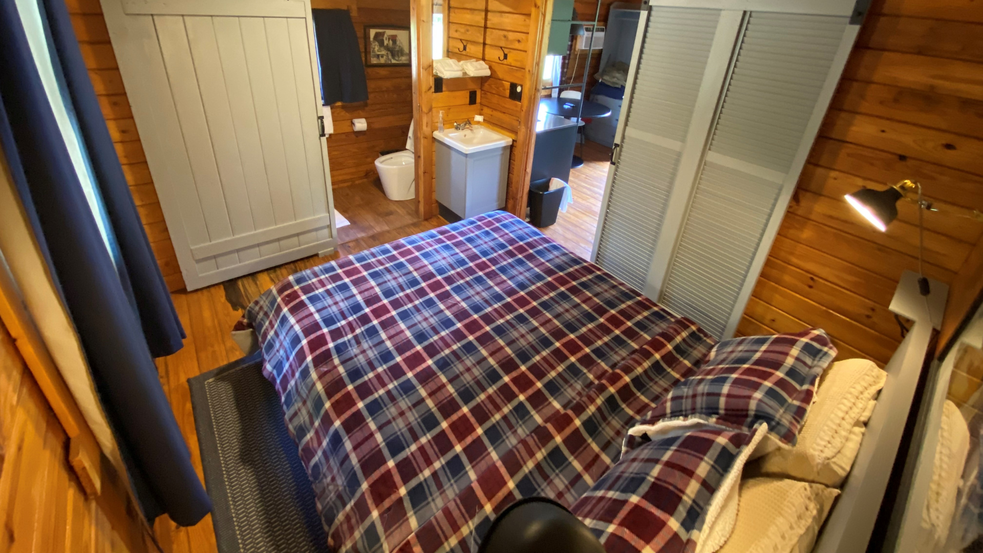 The interior bedroom of a wooden log cabin featuring a plaid bedspread, sliding barn doors, a vanity sink, and a view into the bathroom.