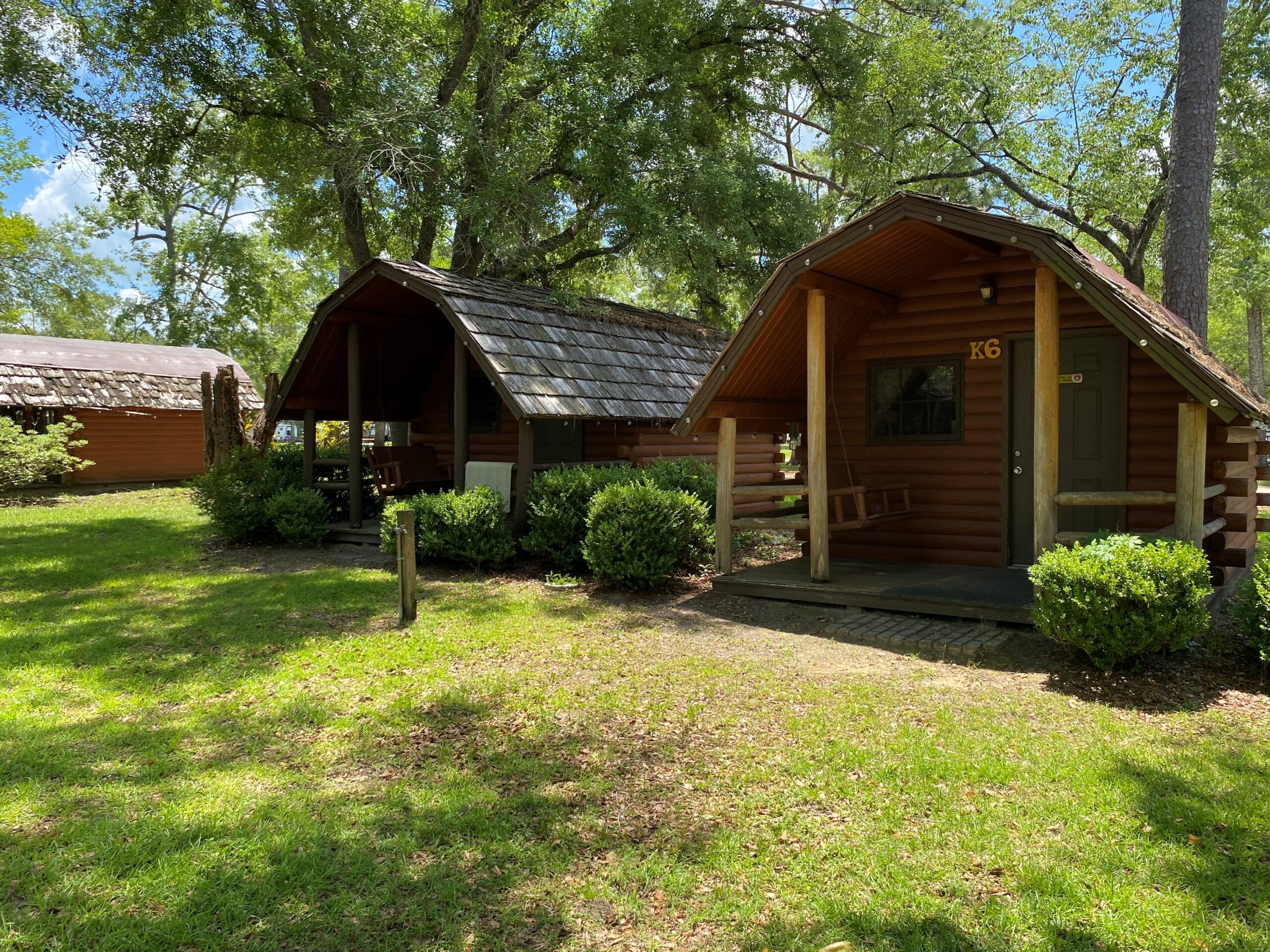 Two rustic wooden log cabins with front porches, porch swings, and shingled roofs in a shaded grassy area at Tallahassee East RV Park.