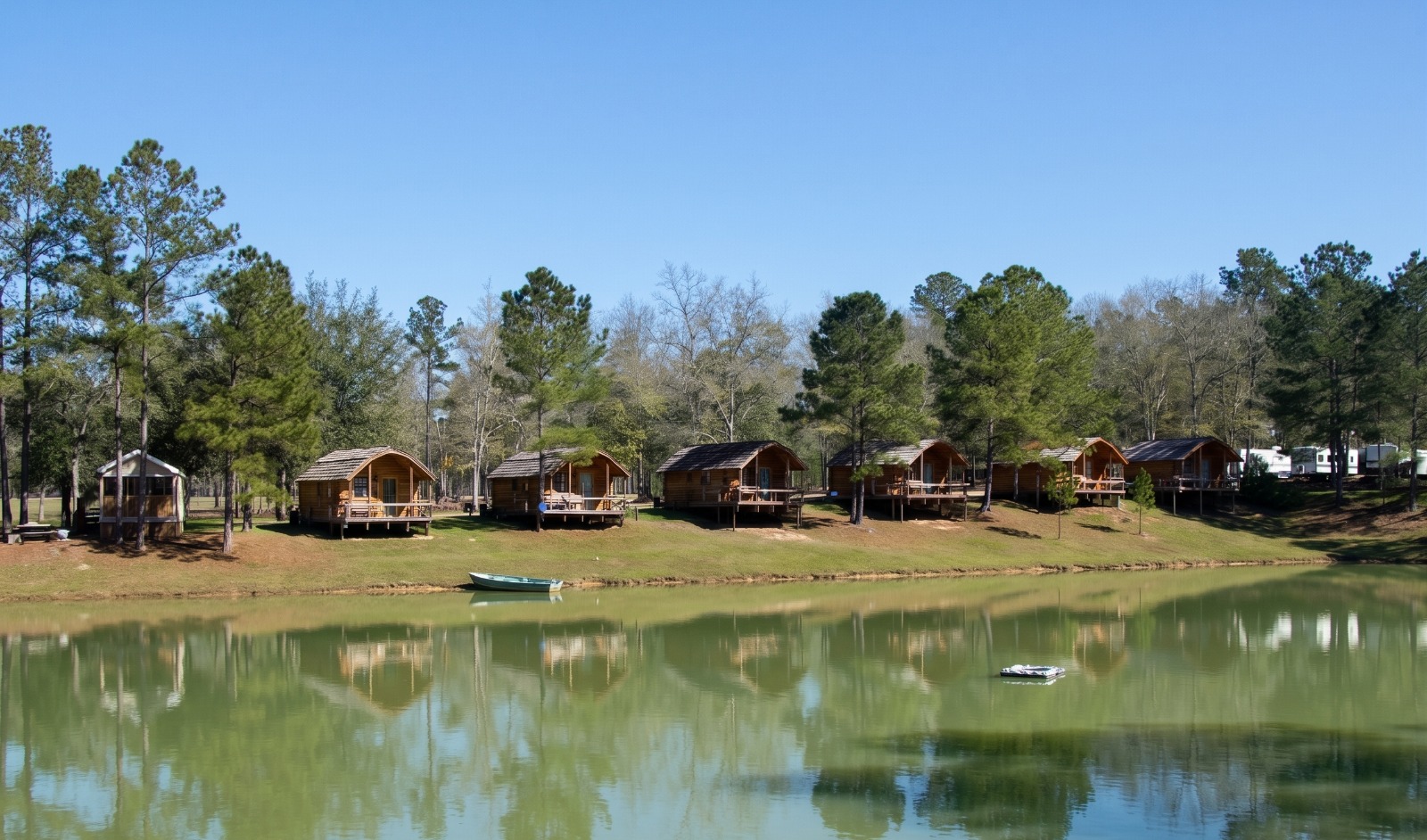 A scenic line of wooden camping cabins with front porches positioned on a grassy hill overlooking a calm green lake.